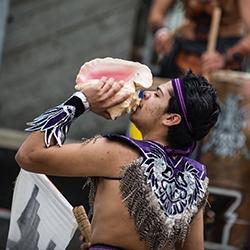 SF State student dressed in purple feathered garment blowing a pink conch